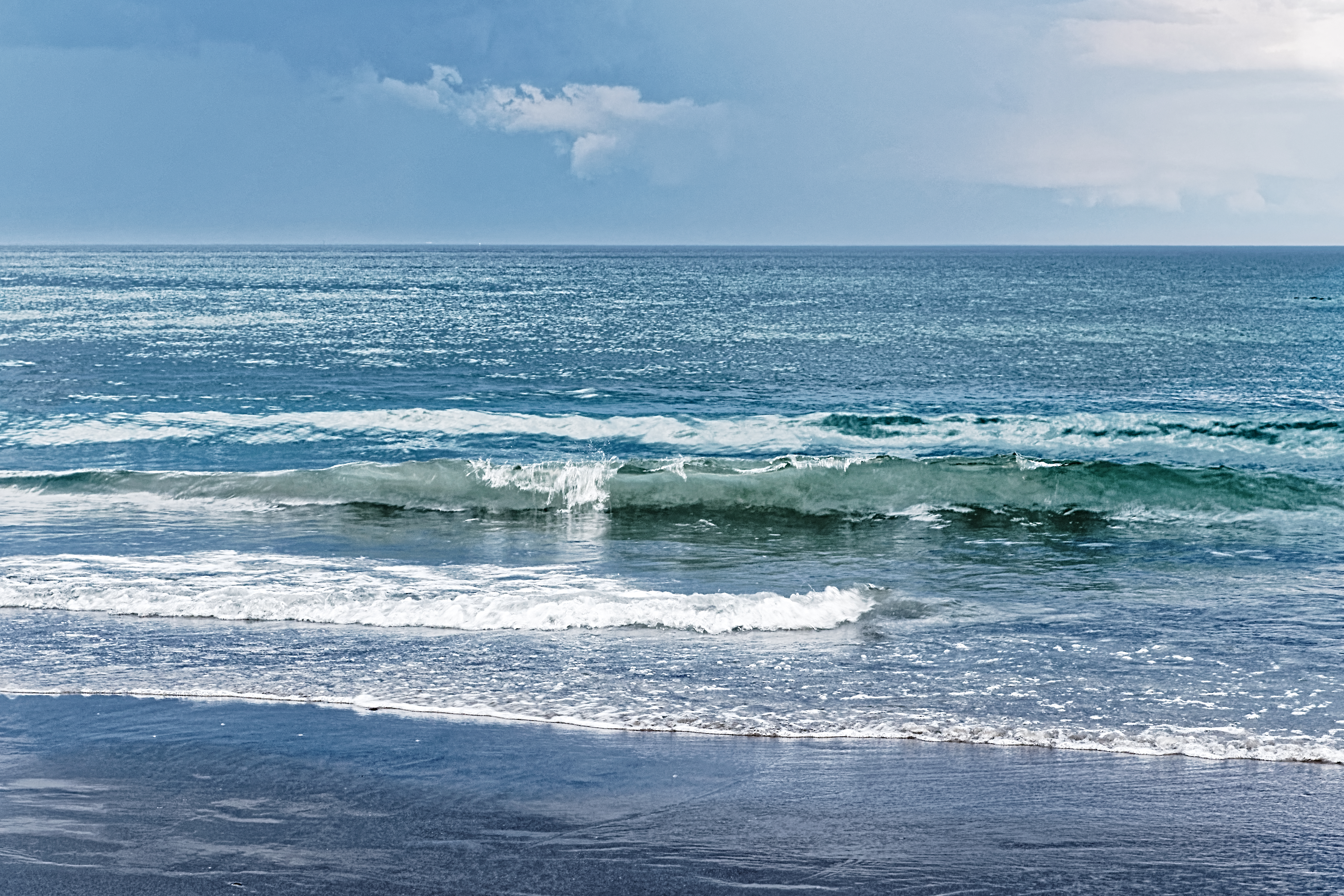 Mostly blue photo of low surf waves with frothy whitecaps coming from a large expanse of ocean and looming, low clouds above with a single white wispy cloud standing out in the center of the sky portion