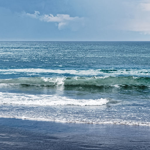 Mostly blue photo of low surf waves with frothy whitecaps coming from a large expanse of ocean and looming, low clouds above with a single white wispy cloud standing out in the center of the sky portion