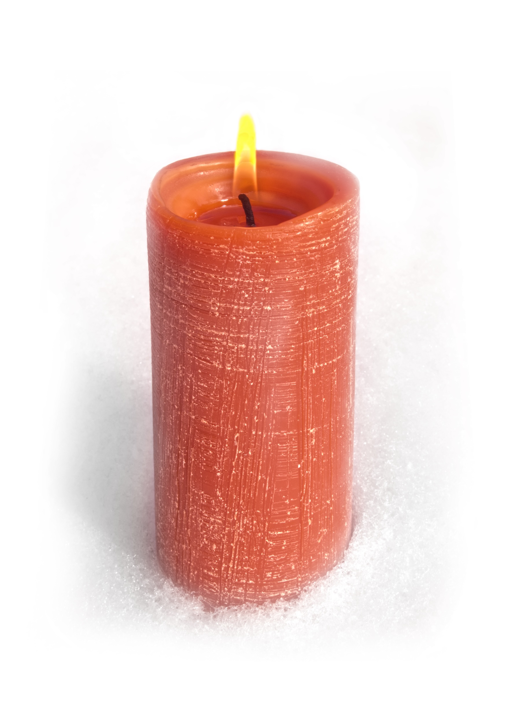 Photo of a lit red candle with a yellow flame and textured sides showing fine horizontal and vertical lines that have a bit of white coloring to accentuate the textured appearance, set onto what appears to be a snow-covered surface