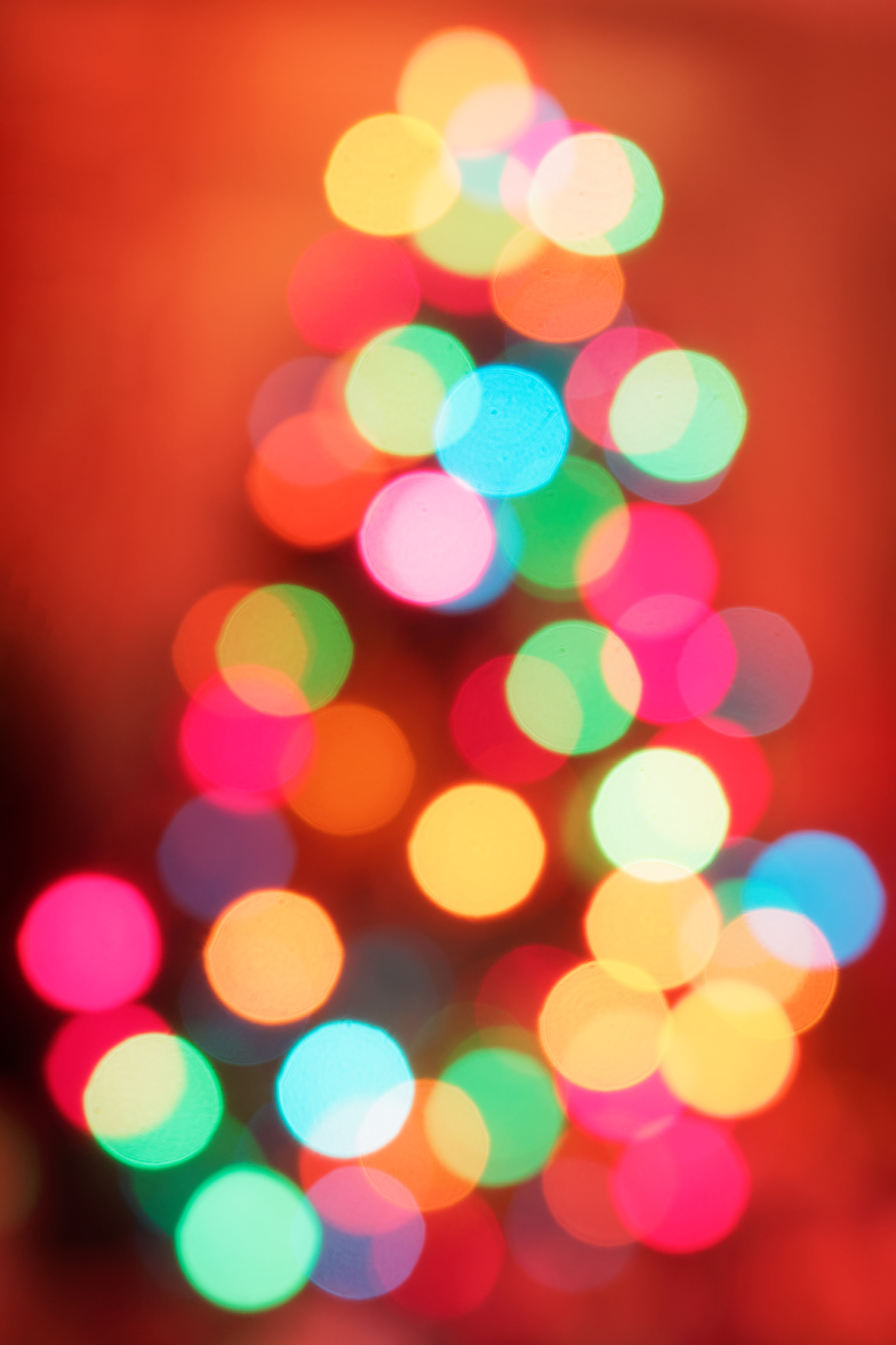 Abstract photo of what appears to be a tree decorated with colored lights but shown so out of focus that the tree itself is not visible other than in form and the lights are bright luminous balls of yellows, reds, oranges, blues, and greens, all with a mottled red background