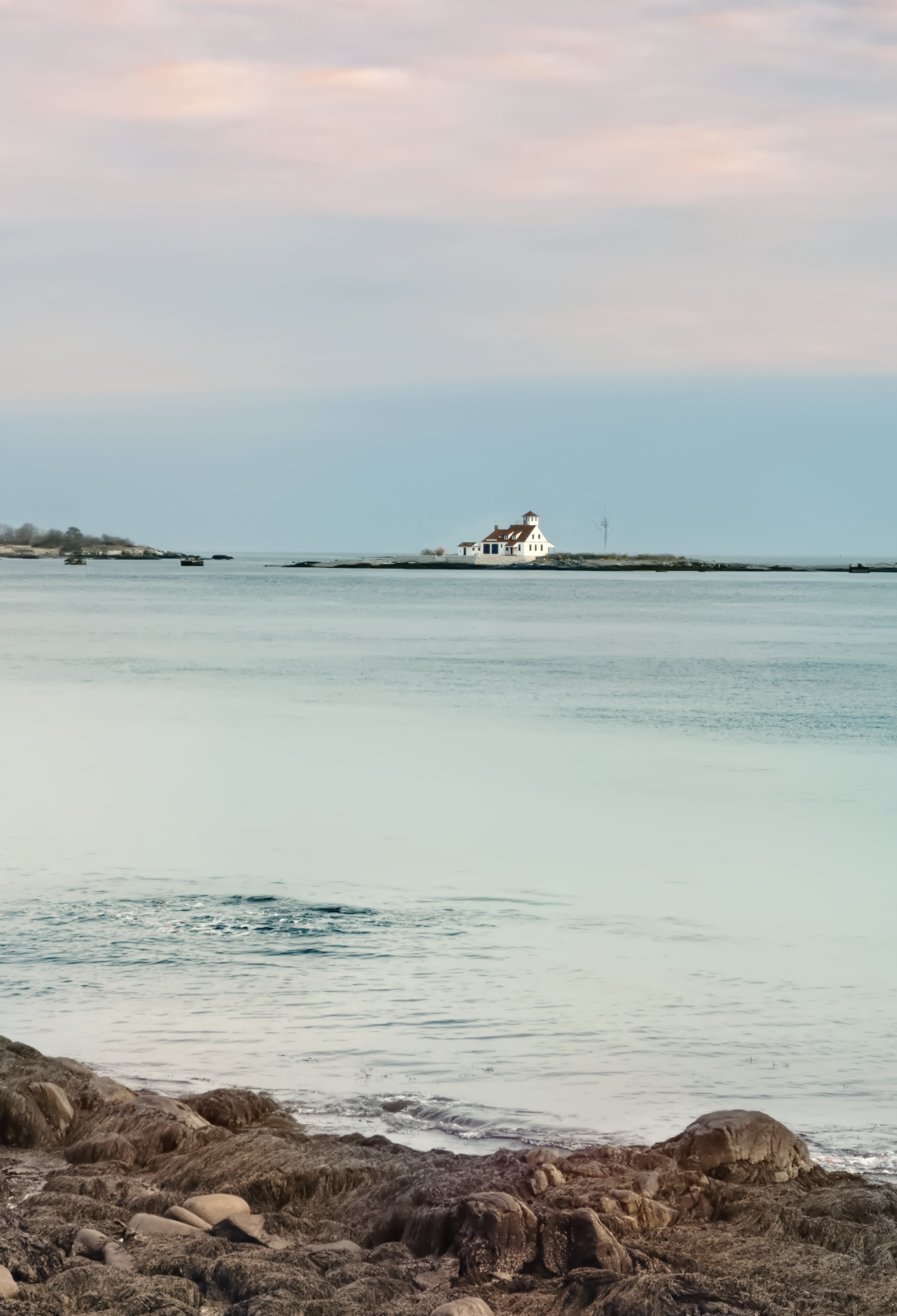 Photo that shows a view, across a calm seafront of a white house in the distance with a square-shaped cupola on top backed by an evening sky of gray-blue and red-tinted clouds