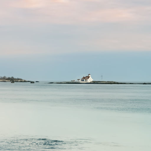 Photo that shows a view, across a calm seafront of a white house in the distance with a square-shaped cupola on top backed by an evening sky of gray-blue and red-tinted clouds