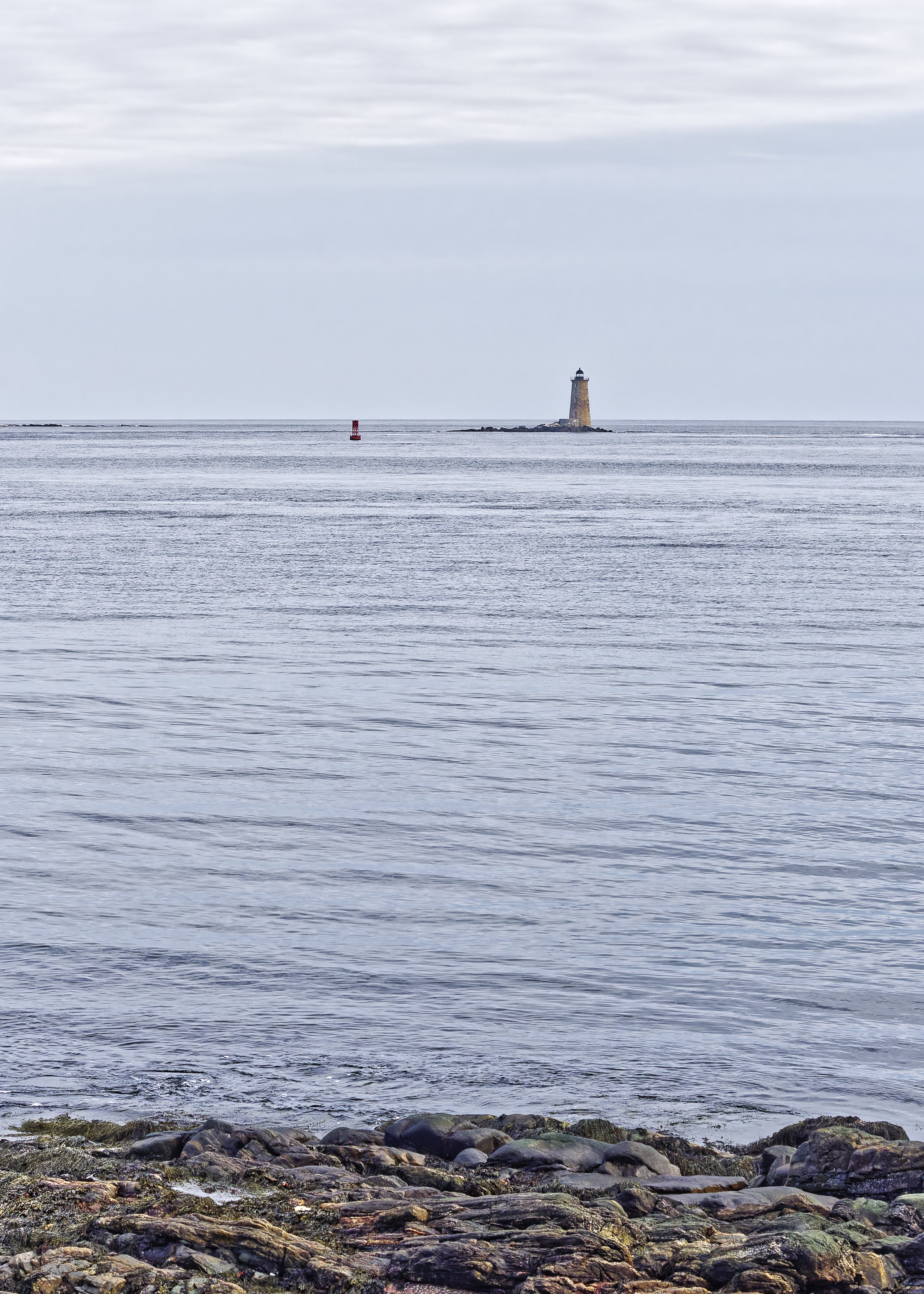 Photo of a stone lighthouse on a very small, rocky island and a nearby floating buoy set in a calm sea with a grayish overcast sky above