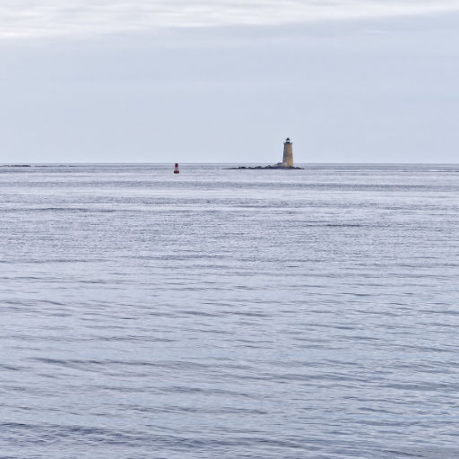 Photo of a stone lighthouse on a very small, rocky island and a nearby floating buoy set in a calm sea with a grayish overcast sky above